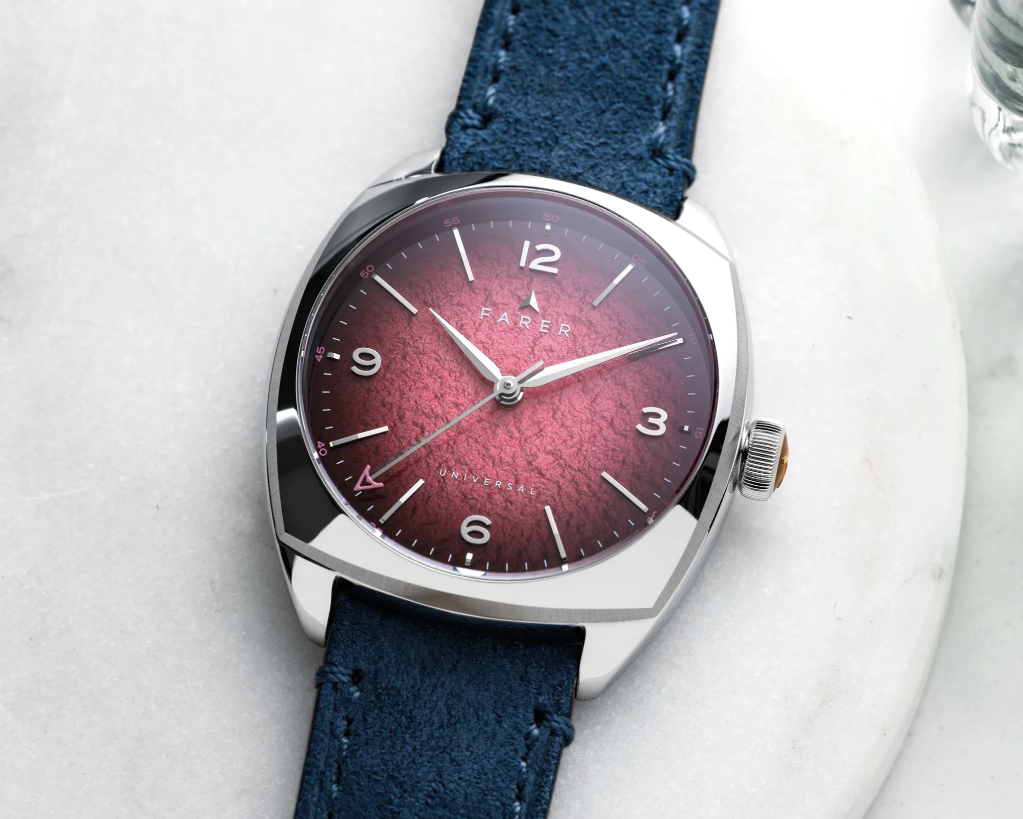 Silver Farer wristwatch with a textured burgundy dial and blue suede strap on a white marble surface.