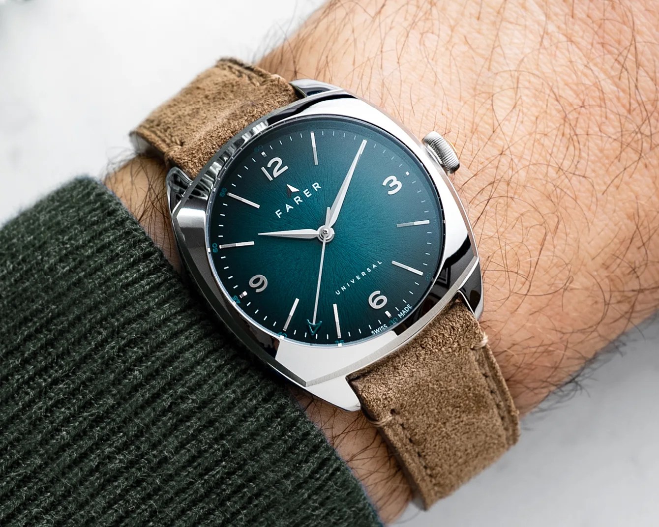 Wristwatch with a teal dial, silver case, and tan suede strap worn on a wrist with a green knitted sleeve.