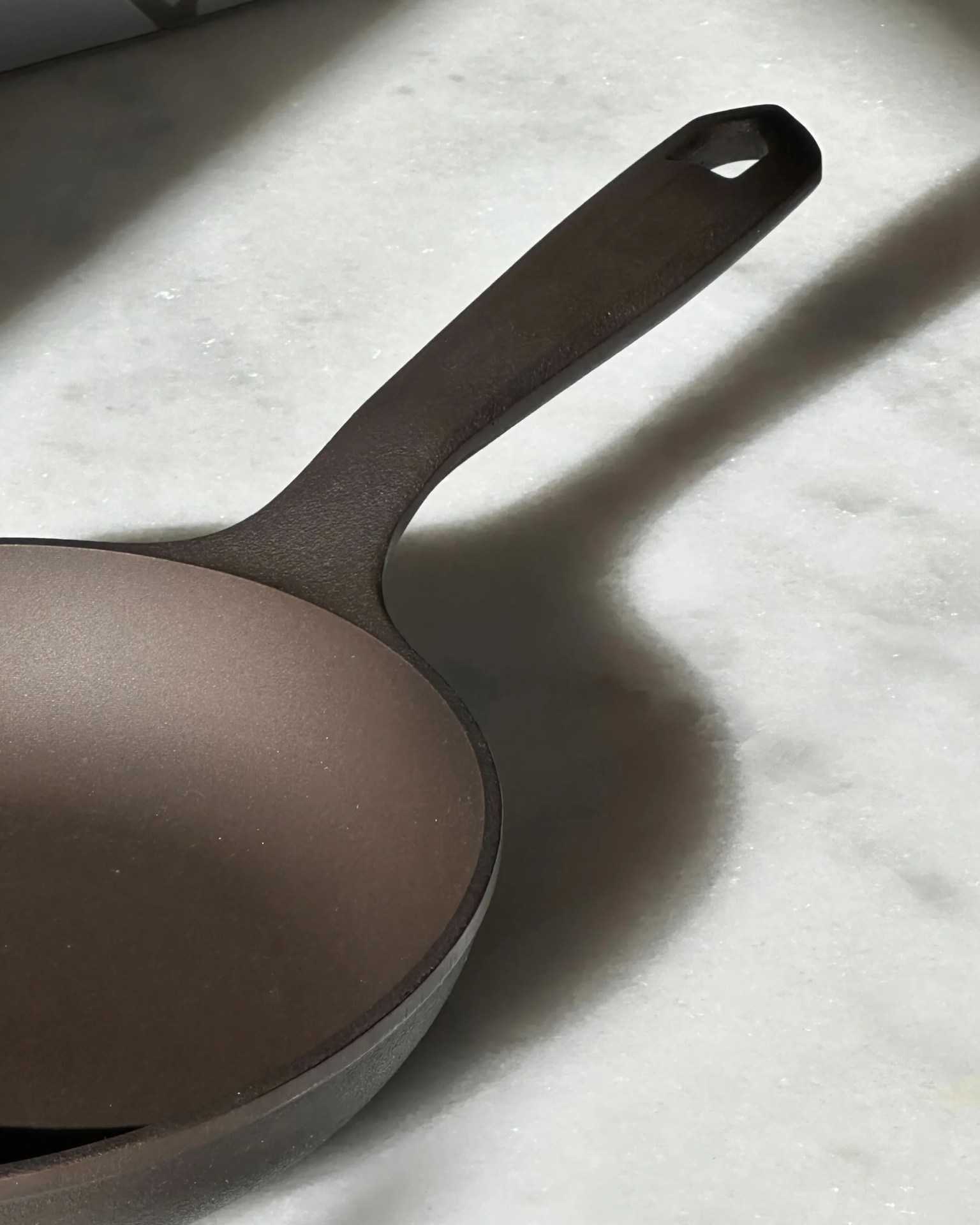 Dark brown cast iron skillet with a handle featuring a hole, placed on a light marble surface.