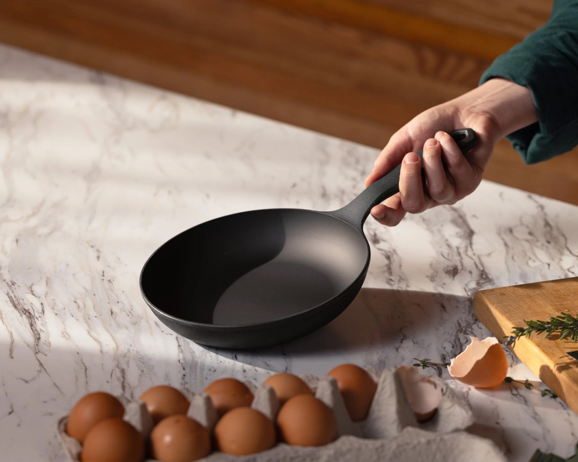 Hand holding a black nonstick frying pan over a marble countertop with brown eggs and cracked eggshells nearby.