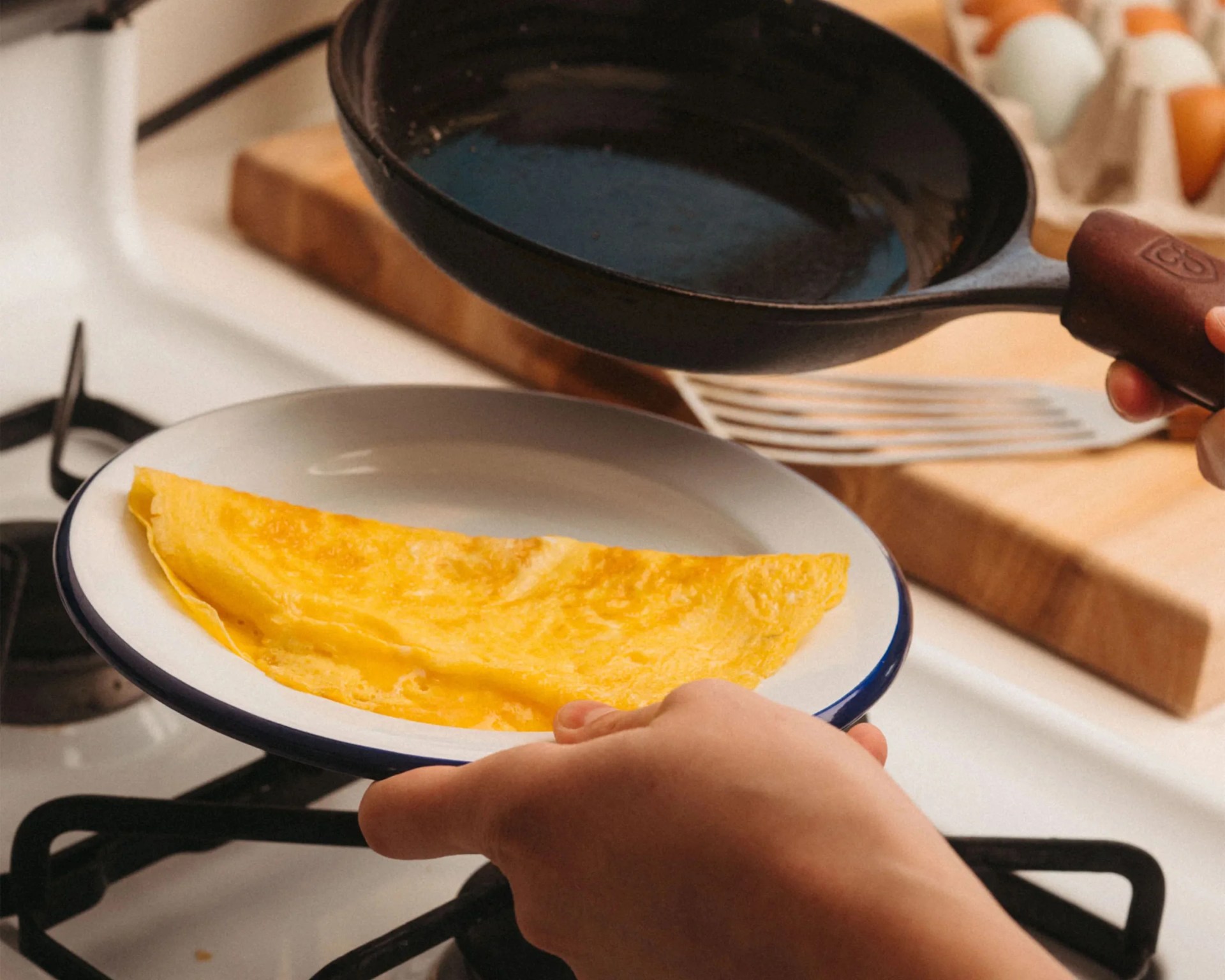 Yellow folded omelette on a white plate with a blue rim held over a gas stove next to a black frying pan.