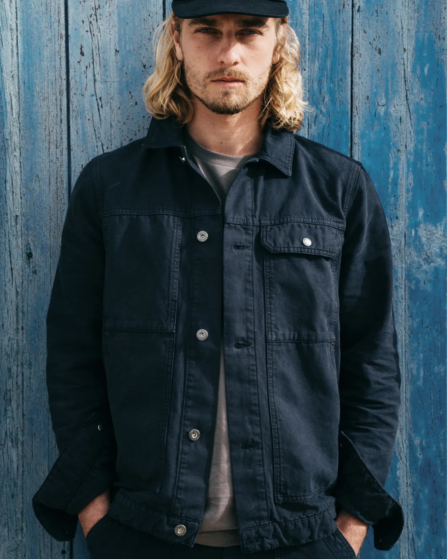Black denim jacket with silver buttons worn by a person standing against a blue wooden wall.