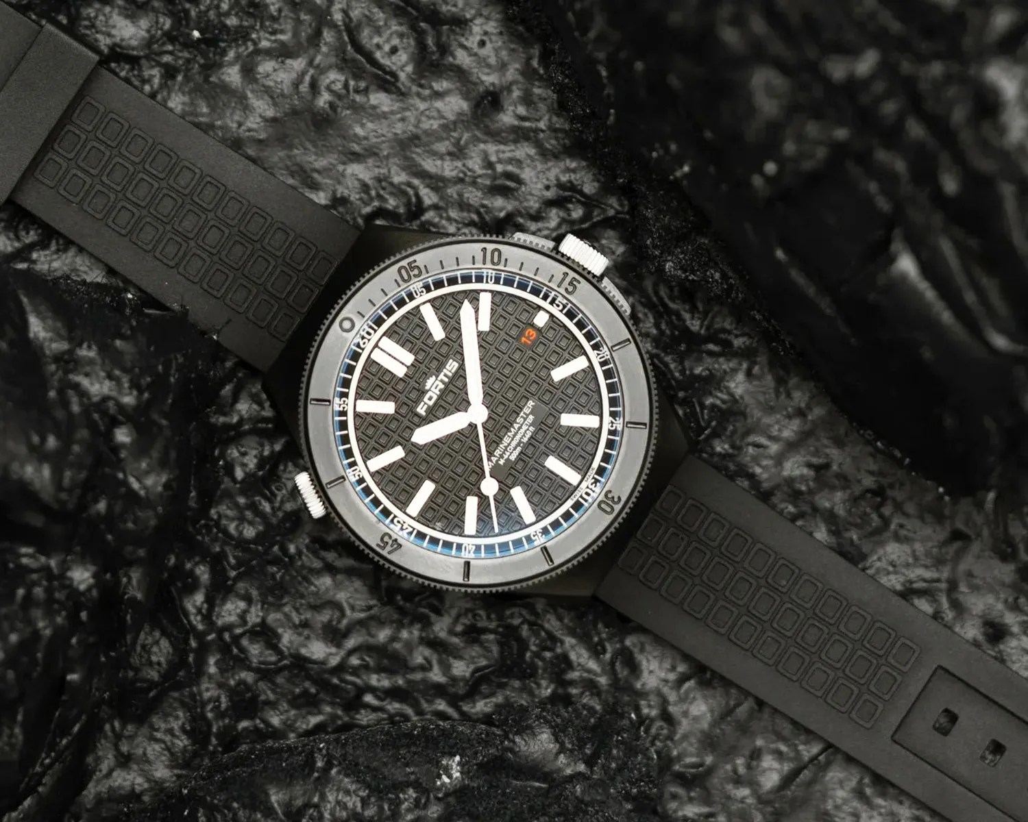 Black Fortis wristwatch with textured dial, white hour markers, and black rubber strap on a dark textured surface.