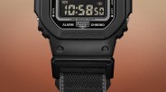 Close-up of a black G-Shock digital watch with LED backlight and black fabric strap against a gradient brown background.
