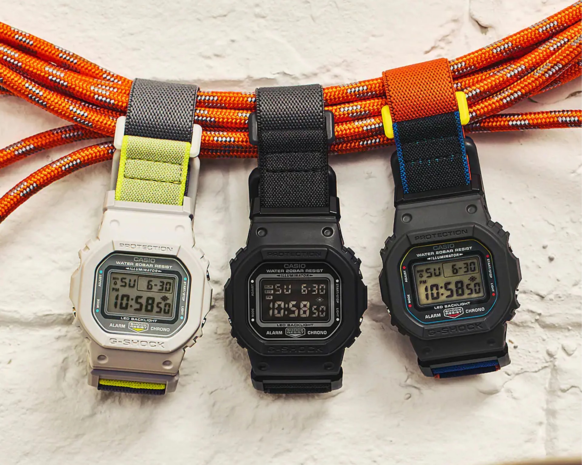 Three Casio G-Shock digital watches with fabric straps in white with yellow, black, and black with orange and blue, hanging on orange climbing ropes.