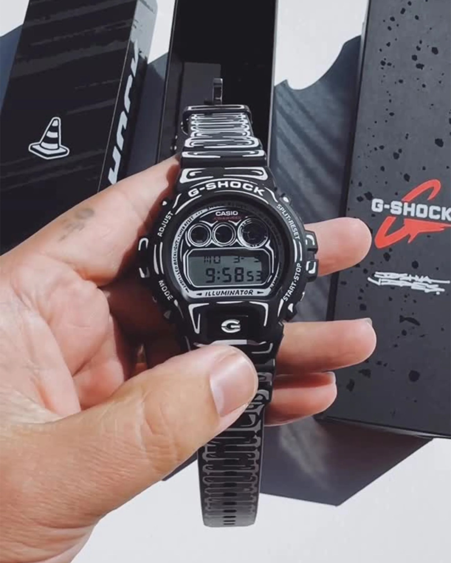 Hand holding a black Casio G-Shock digital watch with white graphic patterns and matching black box in the background.
