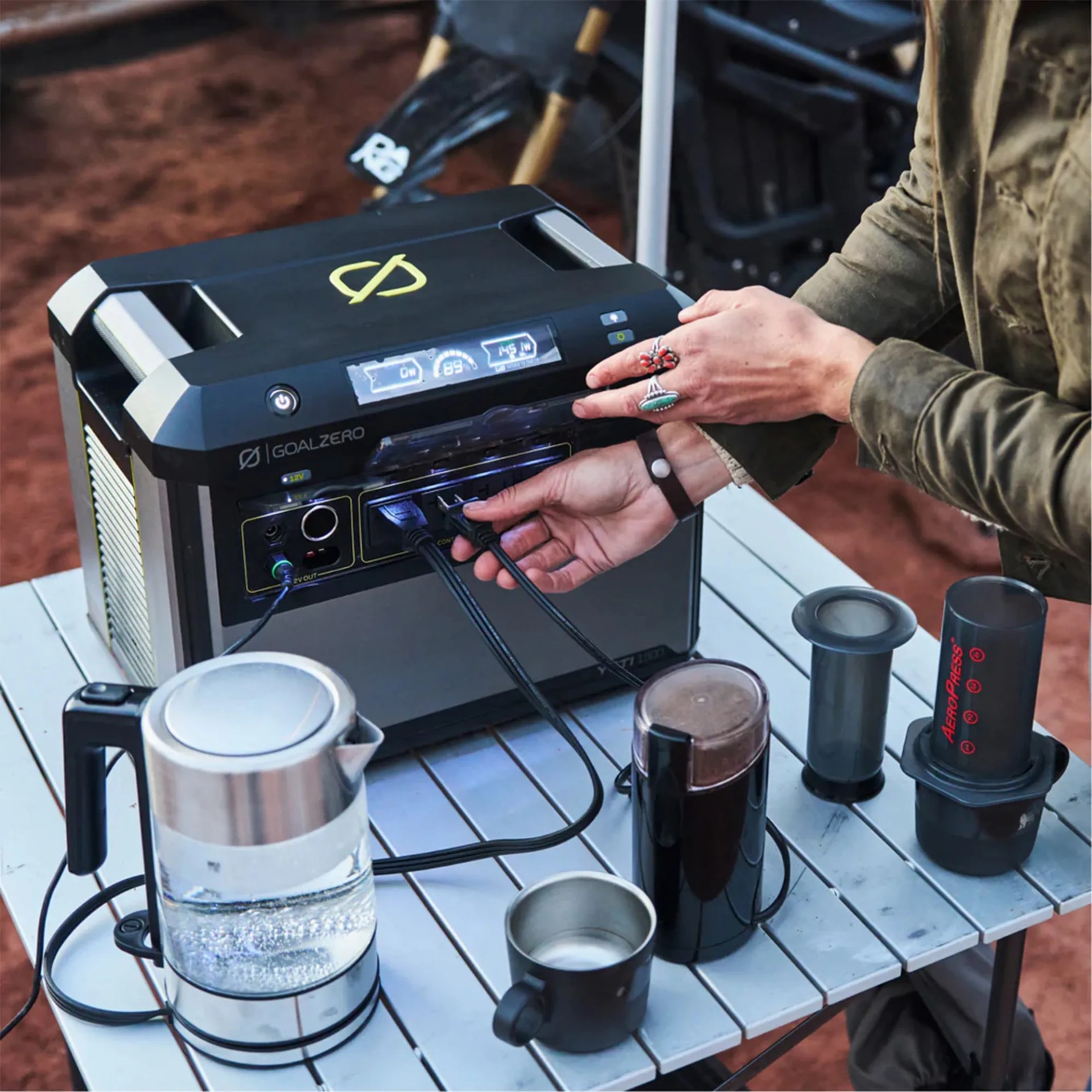 Portable Goal Zero power station with cables connected, powering a kettle, coffee grinder, and AeroPress on a table.