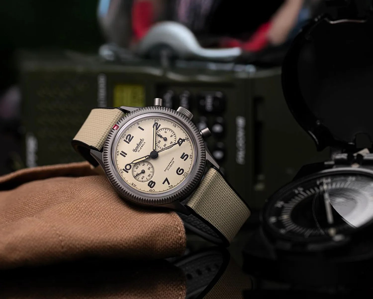 Beige dial wristwatch with black numerals and beige fabric strap resting on brown fabric.