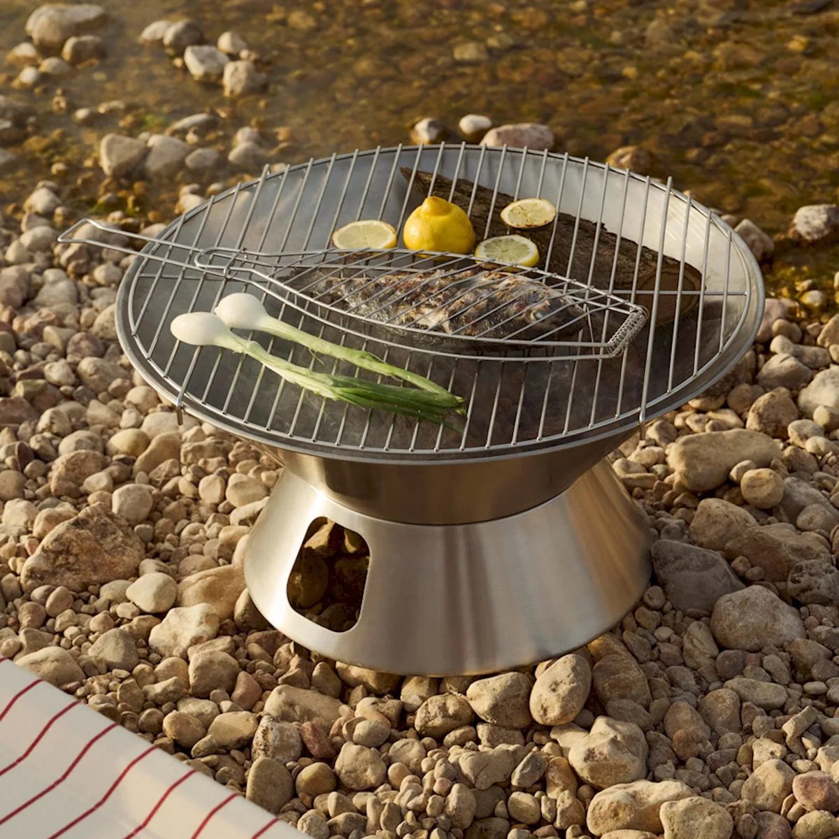 Stainless steel portable grill on rocky ground with fish, lemon slices, and green onions cooking.
