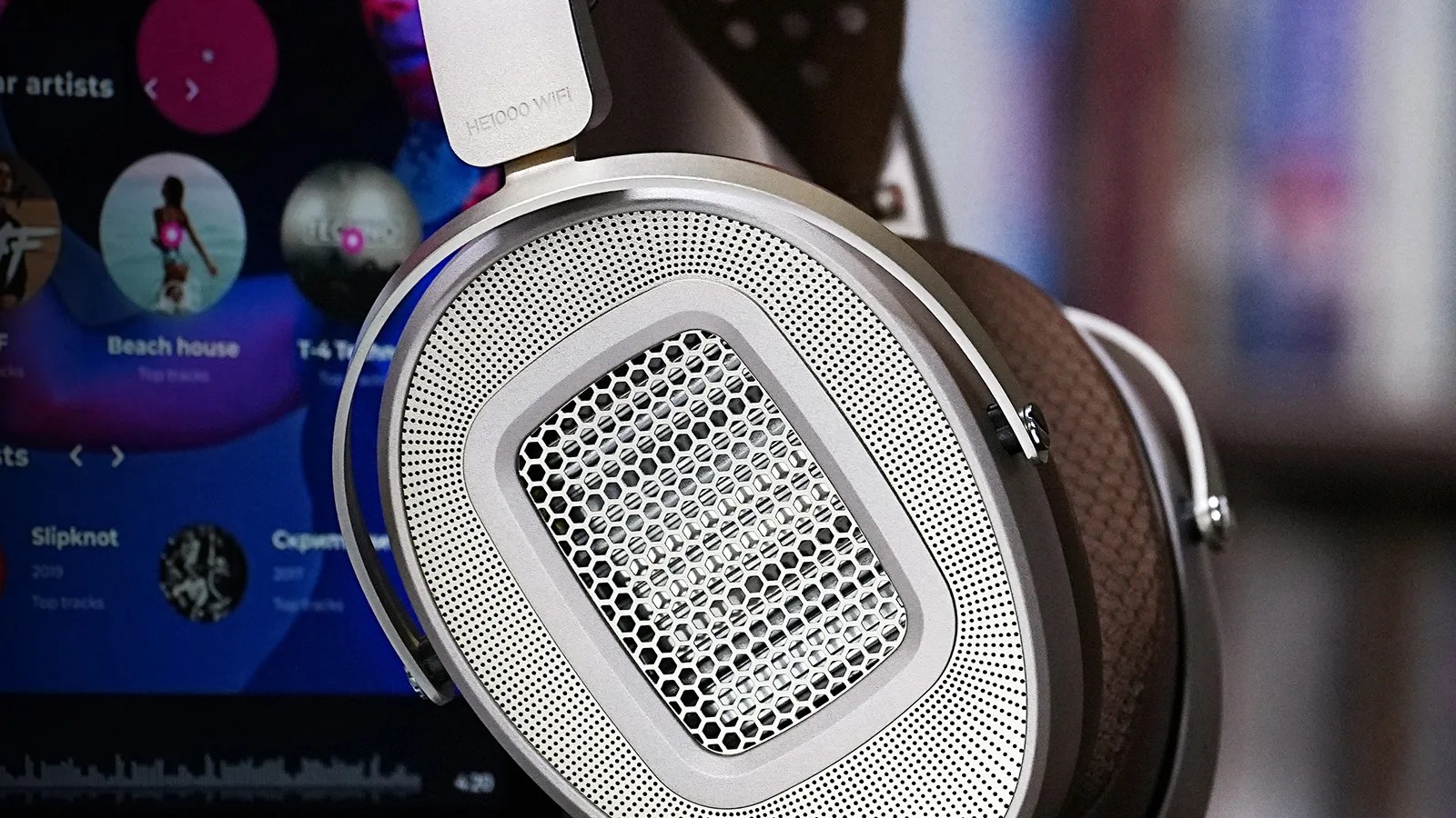 Close-up of silver open-back headphones with hexagonal mesh and brown ear pads in front of a blurred screen.