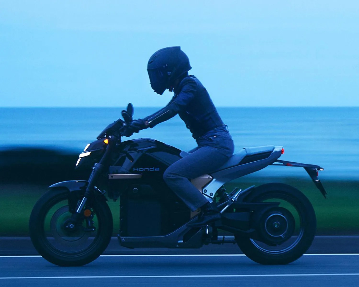 Person wearing black helmet and jacket riding a black Honda electric motorcycle by the ocean at dusk.