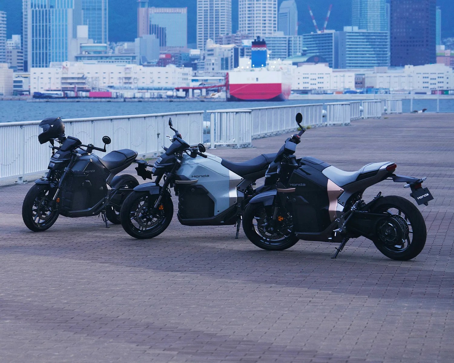 Three Honda electric motorcycles in black and silver parked on a waterfront promenade with a city skyline and ship in the background.