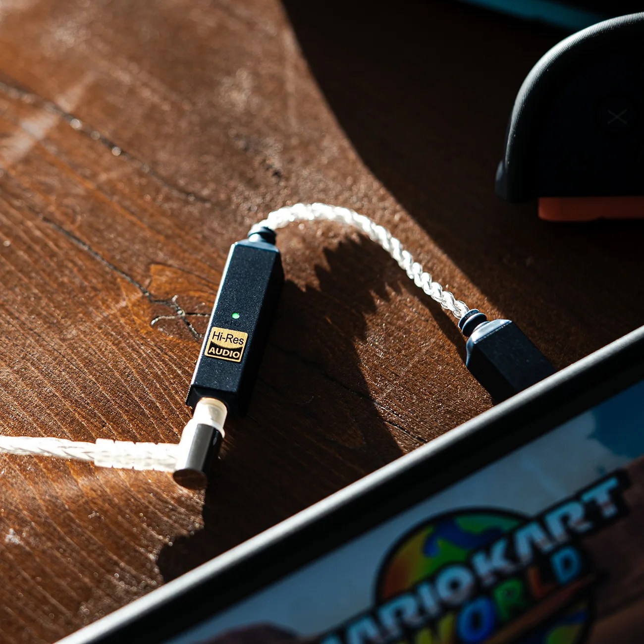 Black Hi-Res Audio adapter with braided cable connected to a device displaying Mario Kart World on screen.
