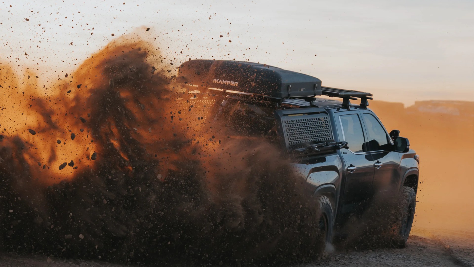 Black off-road vehicle with roof cargo box kicking up dirt and dust while driving on a dusty terrain.