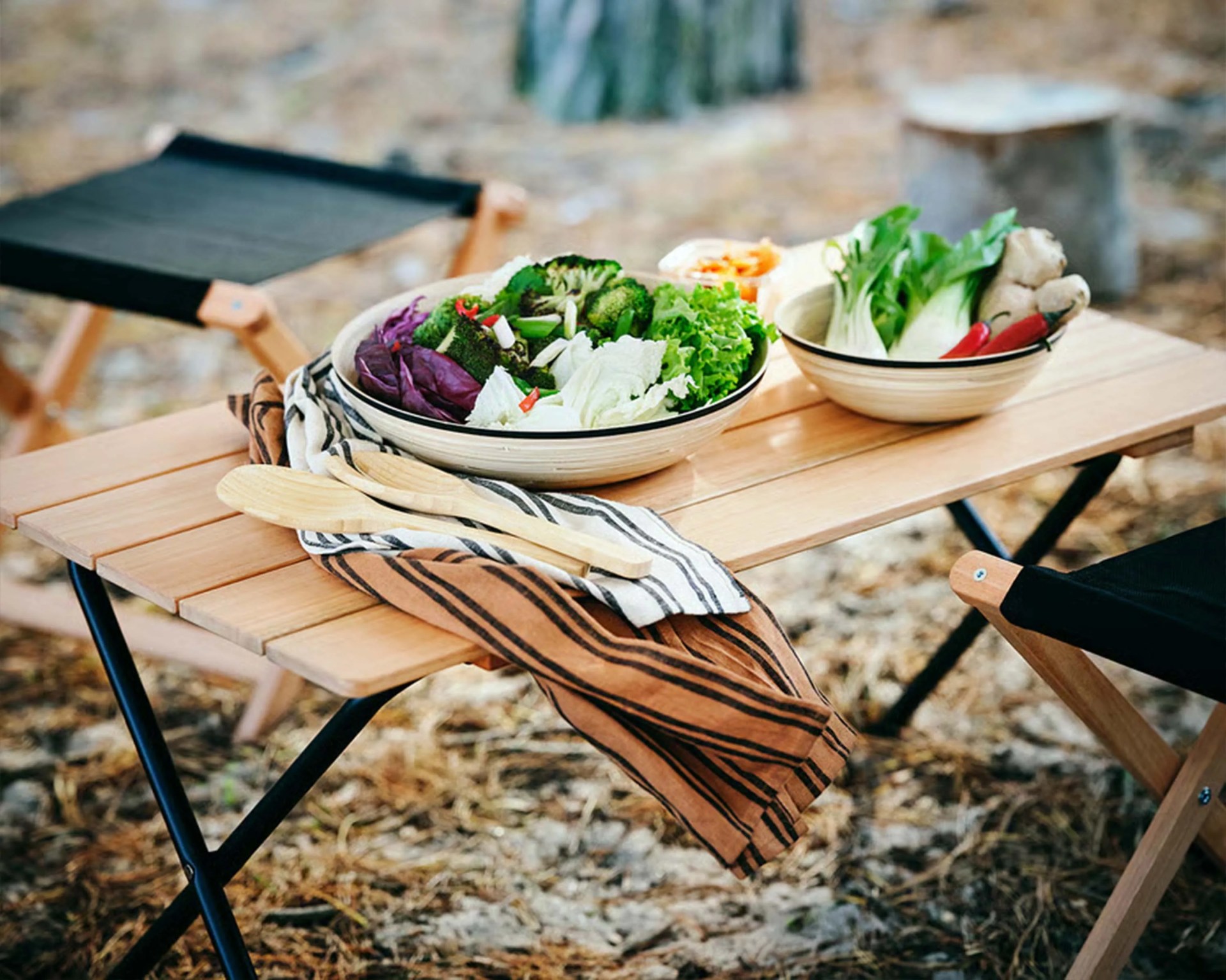 Wooden outdoor picnic table with two bowls of fresh vegetables and striped cloth napkins with wooden utensils.