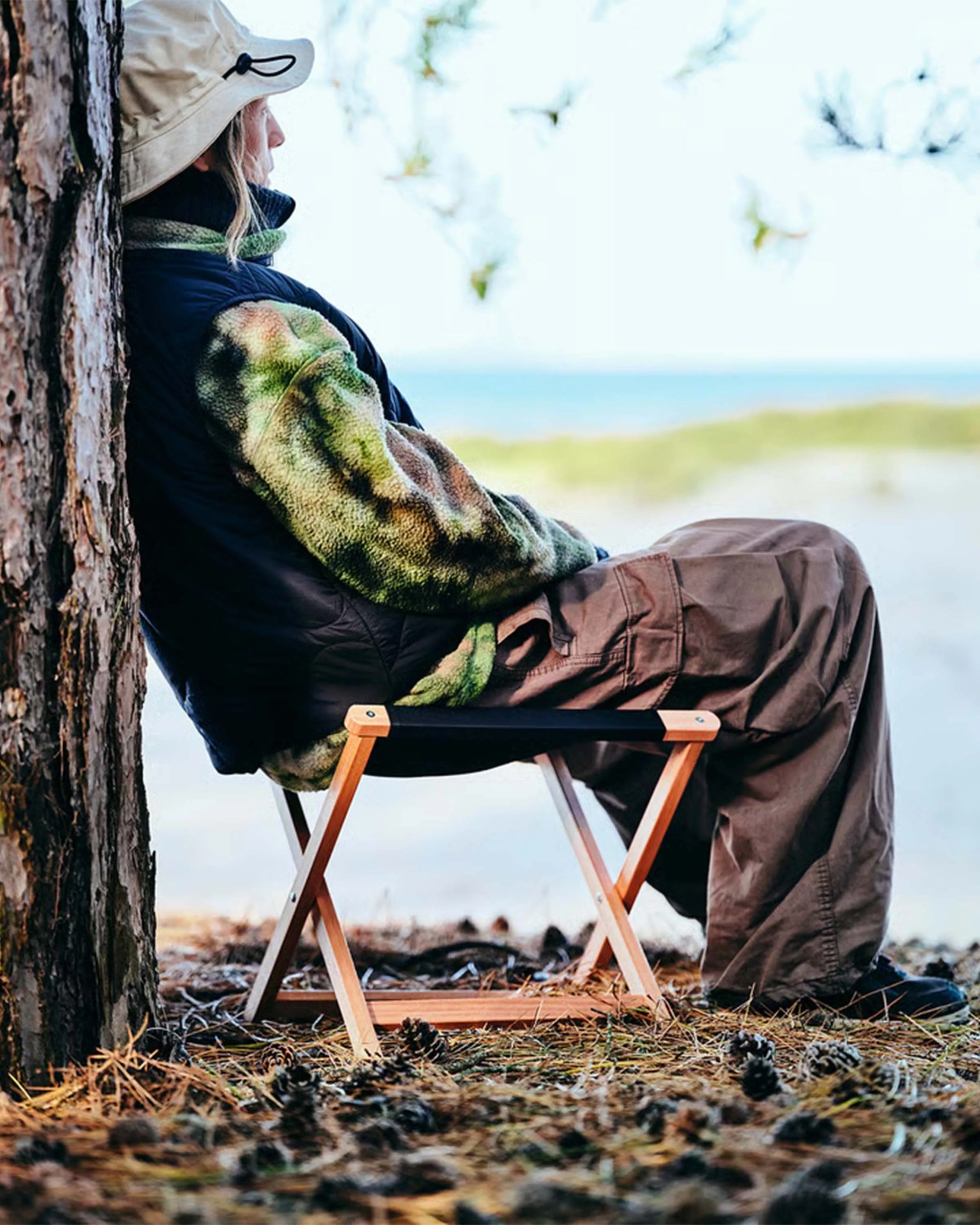 Person wearing a beige bucket hat, green and black fleece jacket, and brown pants sitting on a small wooden folding stool outdoors.