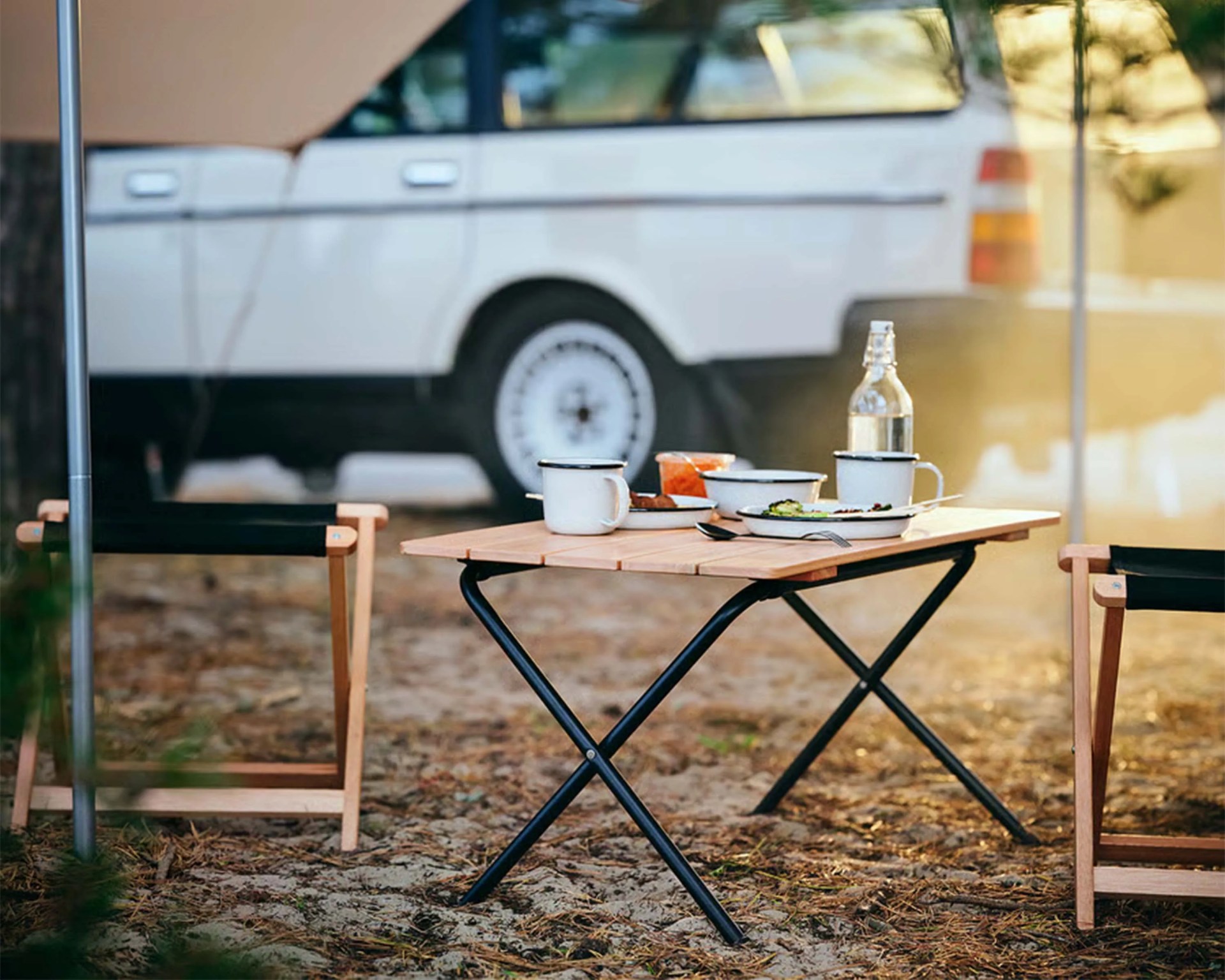 Wooden camping table with black metal legs set with enamel mugs, plates, and a glass bottle, flanked by two wooden chairs.