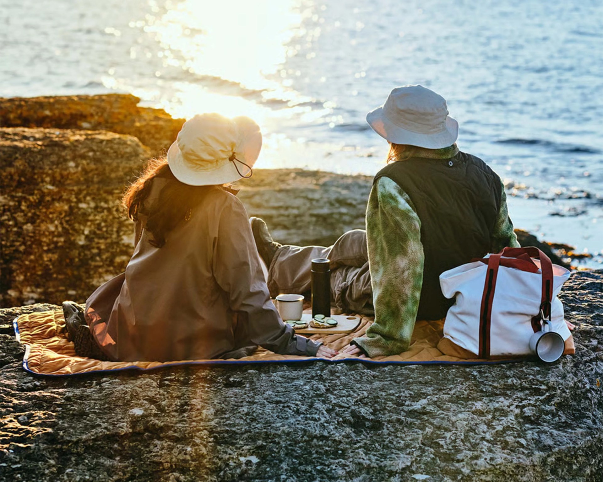 Two people wearing hats sitting on a blanket on rocky ground by the water at sunset with a white bag and picnic items.