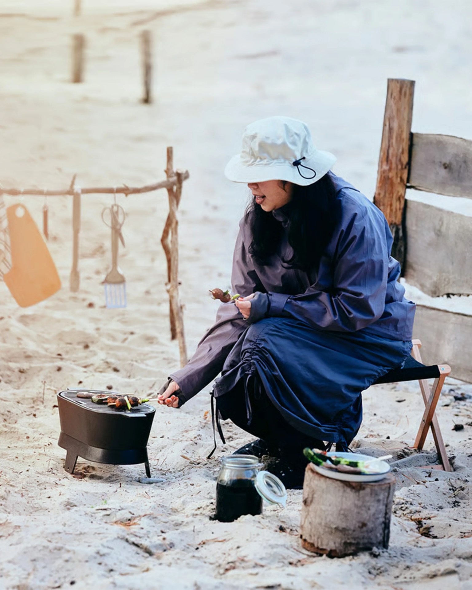 Person wearing a white bucket hat and dark clothing grilling skewers on a small black portable grill on sandy ground.