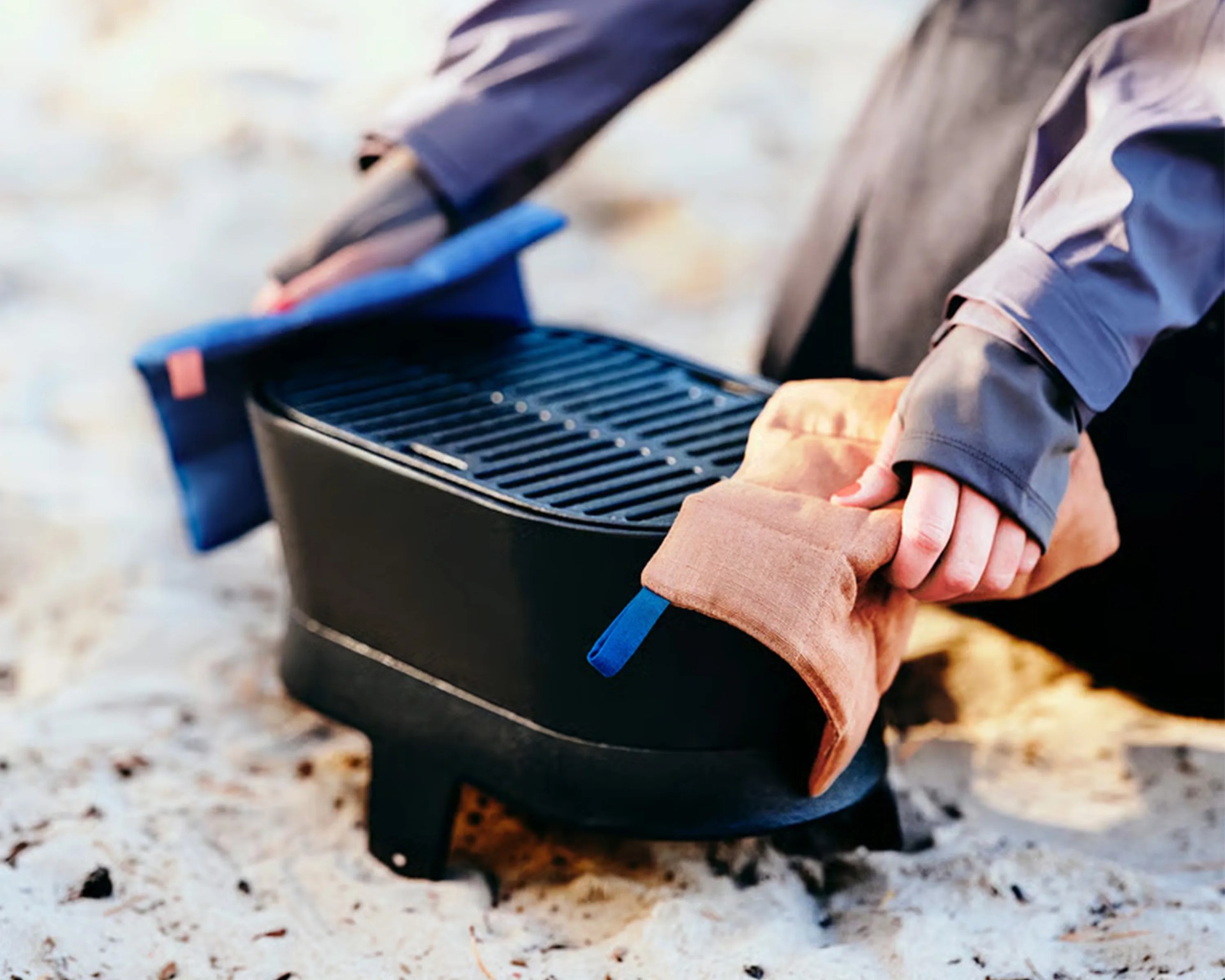 Person using blue and brown heat-resistant gloves to handle a black portable grill on sandy ground.