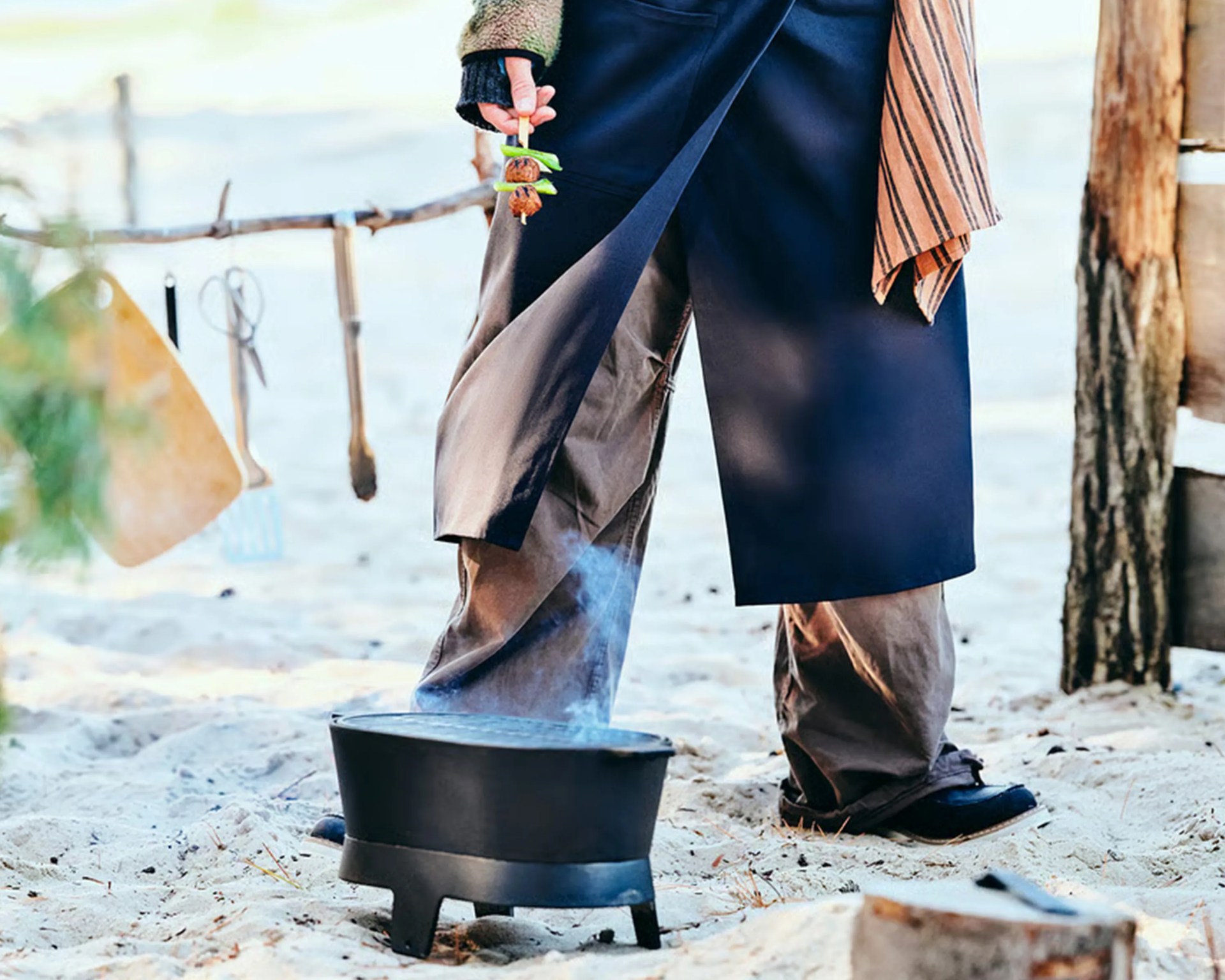 Black cast iron grill with smoke rising on sandy ground near a person holding a skewer with grilled food.