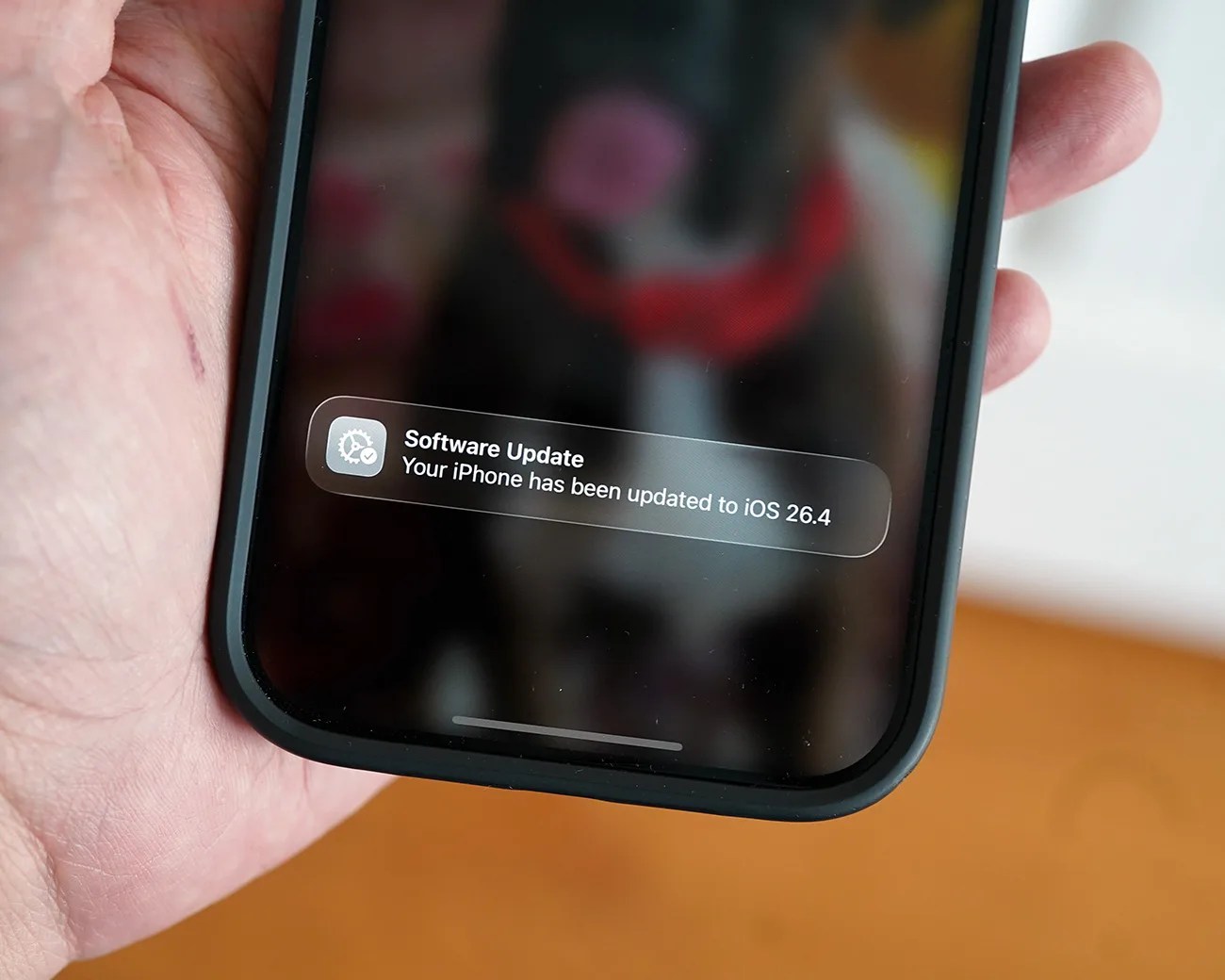 Hand holding a black smartphone displaying a software update notification for iOS 26.4.