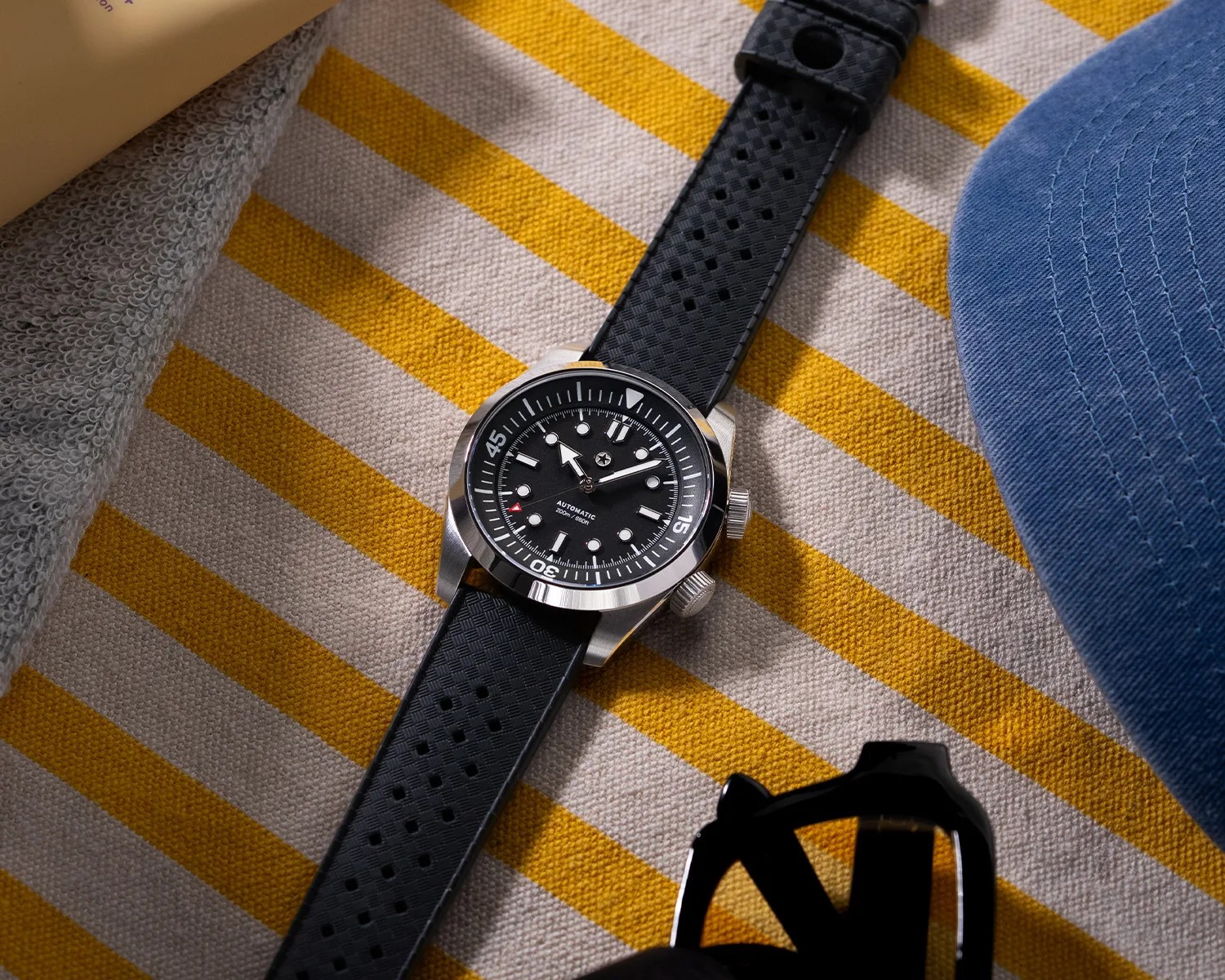Black dive watch with stainless steel case and perforated black rubber strap on yellow and white striped fabric.