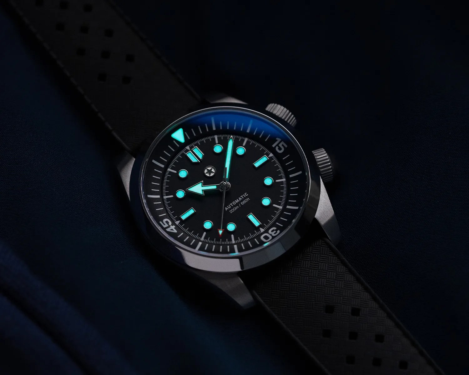 Black dive watch with glowing turquoise hour markers and hands on a black textured rubber strap.