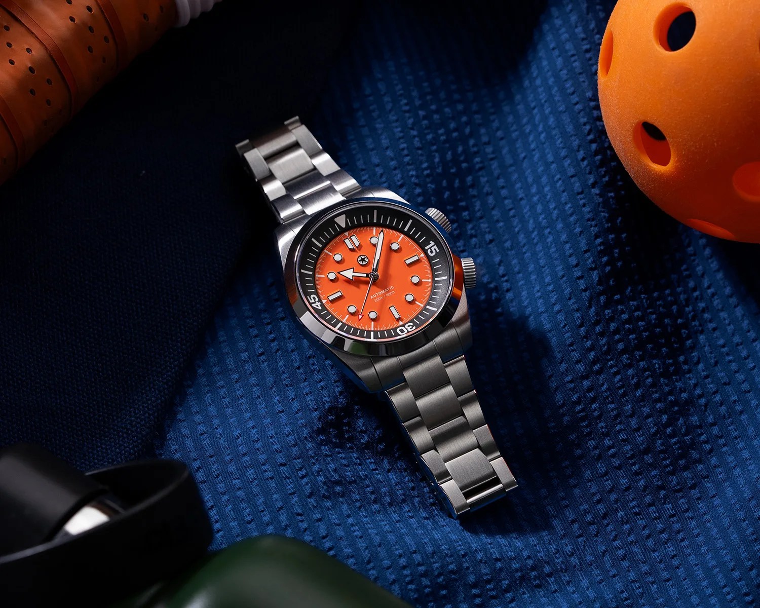 Stainless steel wristwatch with bright orange dial and black bezel on textured blue fabric.