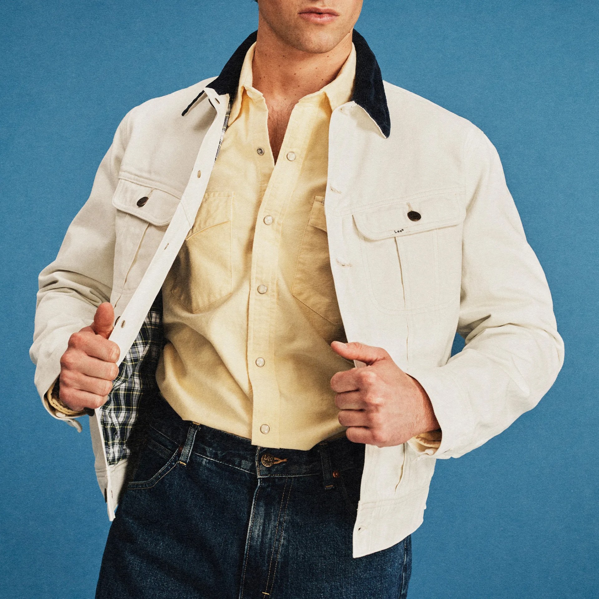 Off-white denim jacket with black collar and plaid lining worn over a pale yellow button-up shirt and dark blue jeans.