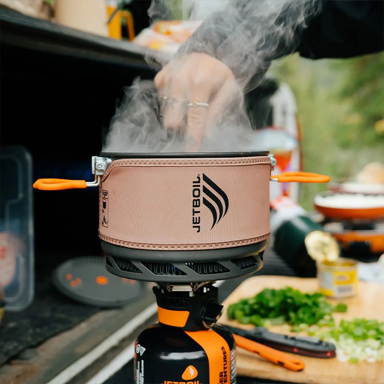 Portable Jetboil camping stove with orange handles and steam rising, set outdoors near a cutting board with chopped greens.