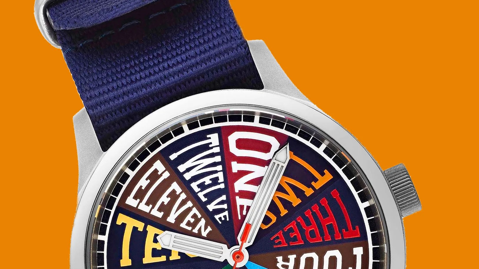 Close-up of a wristwatch with a silver case, navy fabric strap, and colorful dial displaying numbers as words.