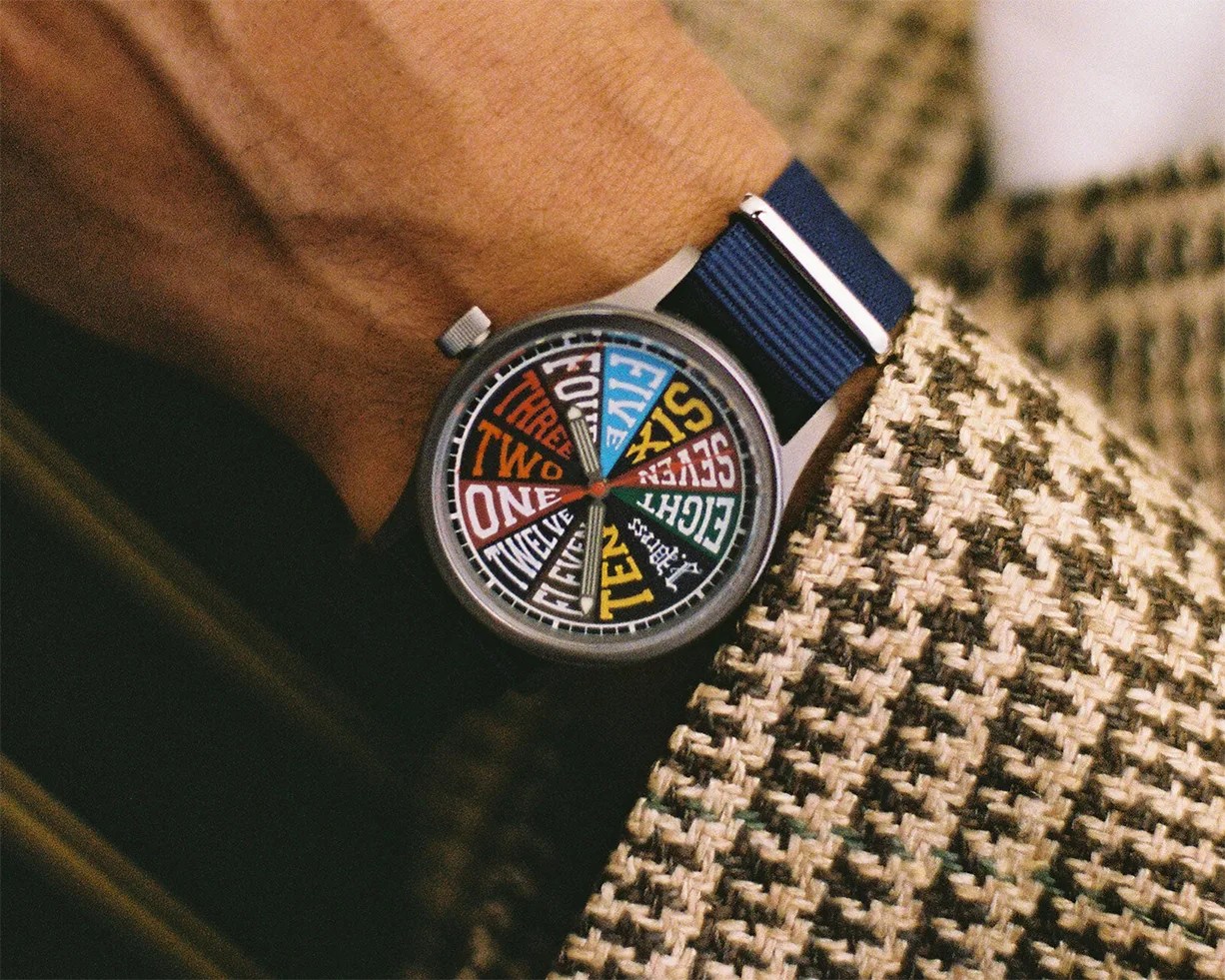 Wristwatch with a colorful dial displaying numbers as words and a blue fabric strap worn with a houndstooth patterned jacket.