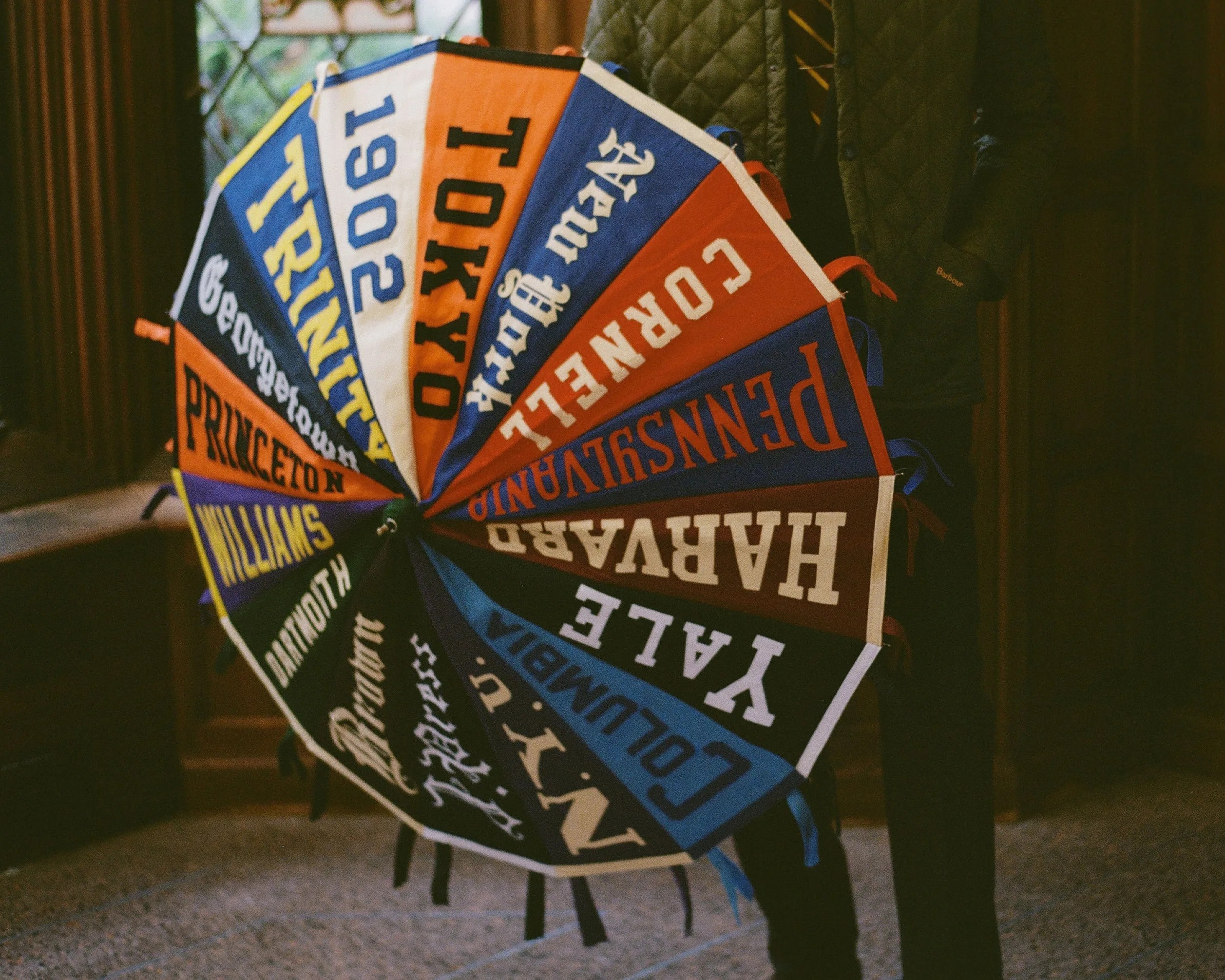 Colorful umbrella with names of universities and locations printed on each panel, held by a person wearing a dark jacket.