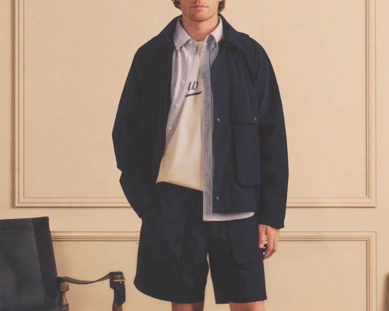 Man wearing a navy jacket and matching shorts over a striped button-up shirt and white graphic sweatshirt.