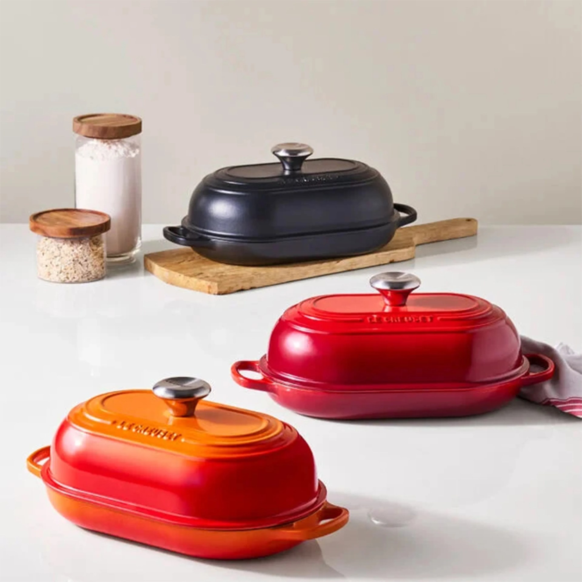 Three oval cast iron covered roasters in black, red, and orange on a white countertop with wooden cutting board and glass jars in the background.
