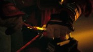 Hand wearing a black wrist strap gripping a red climbing rope attached to a metal carabiner in low light.