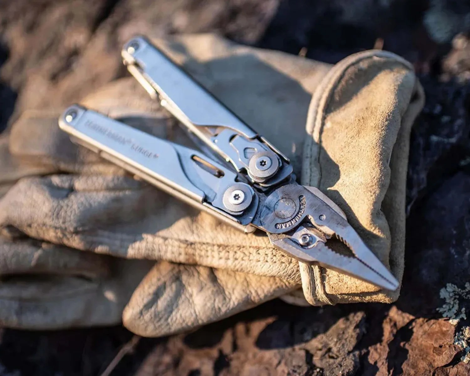 Stainless steel Leatherman multi-tool pliers resting on a pair of worn beige work gloves on rocky ground.