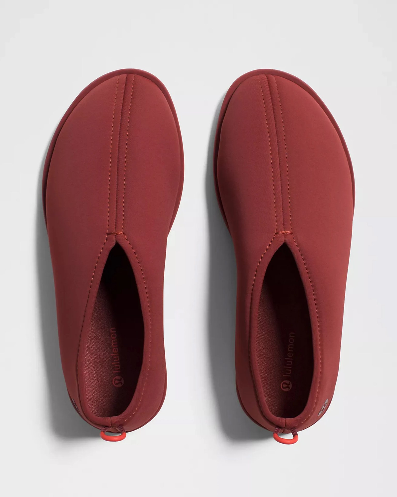 Pair of rust-colored slip-on shoes with rounded toes and small red pull loops at the heel.