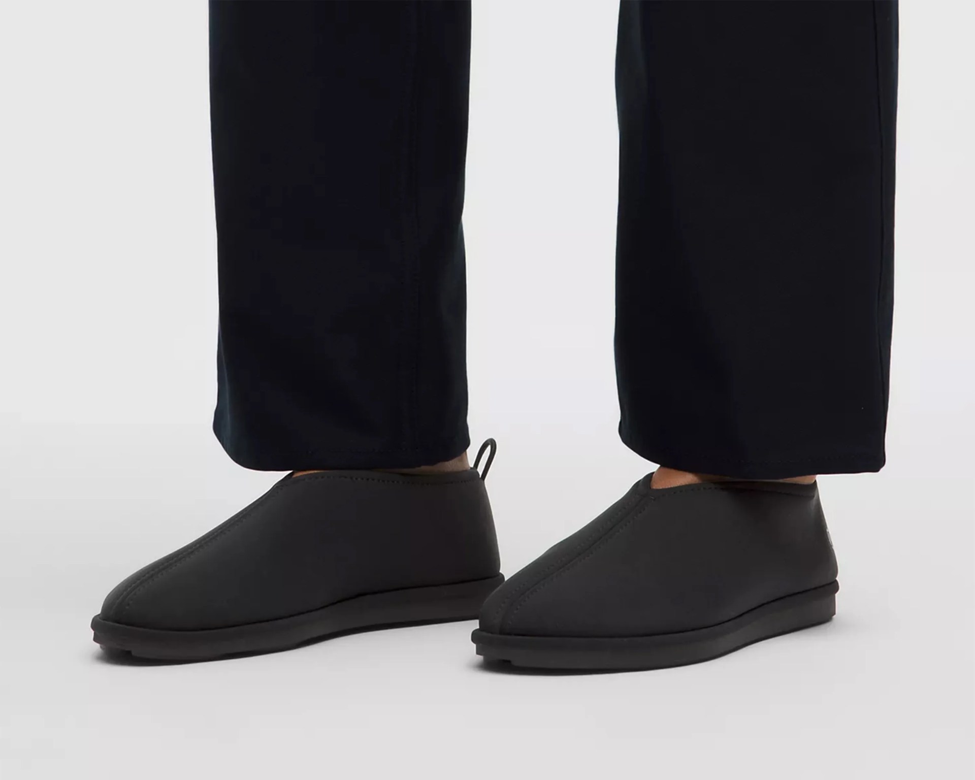 Pair of black slip-on shoes with rounded toes and pull tabs at the back, worn with black pants.