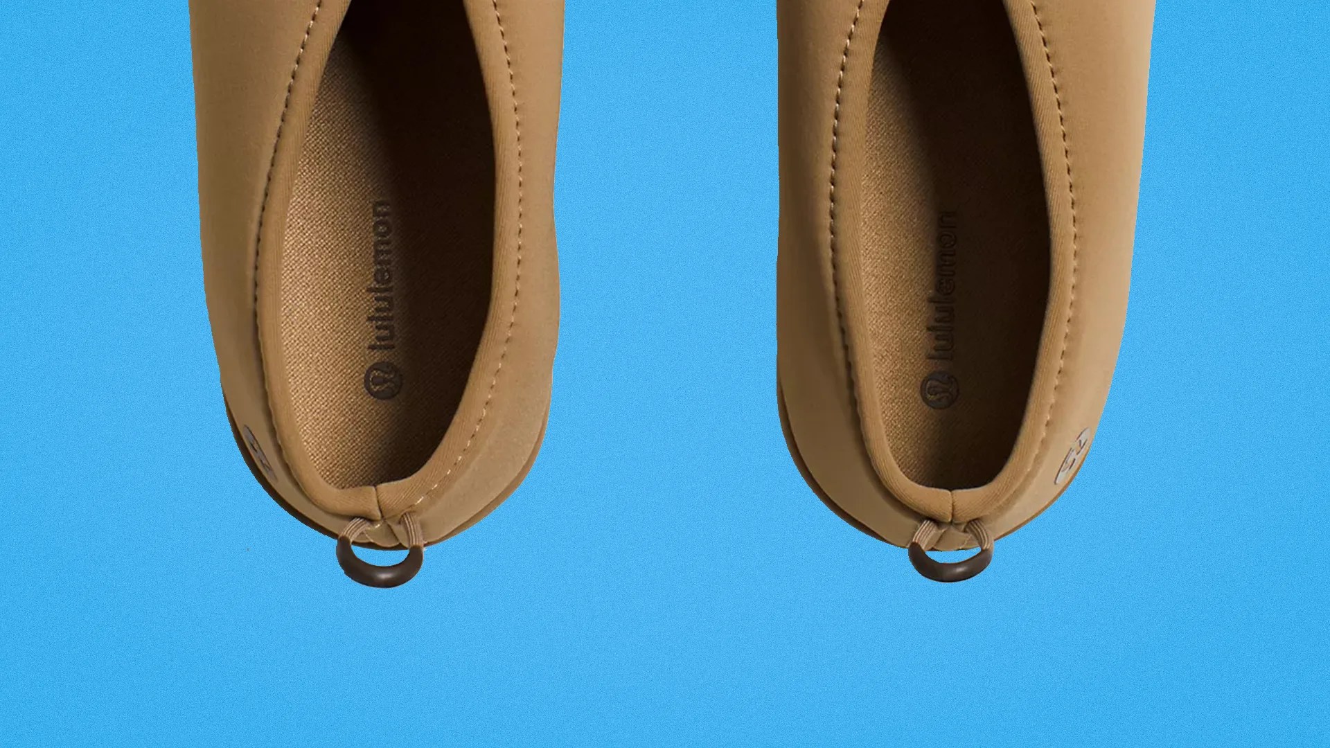 Pair of tan slip-on shoes with fabric lining and pull loops on a blue background.