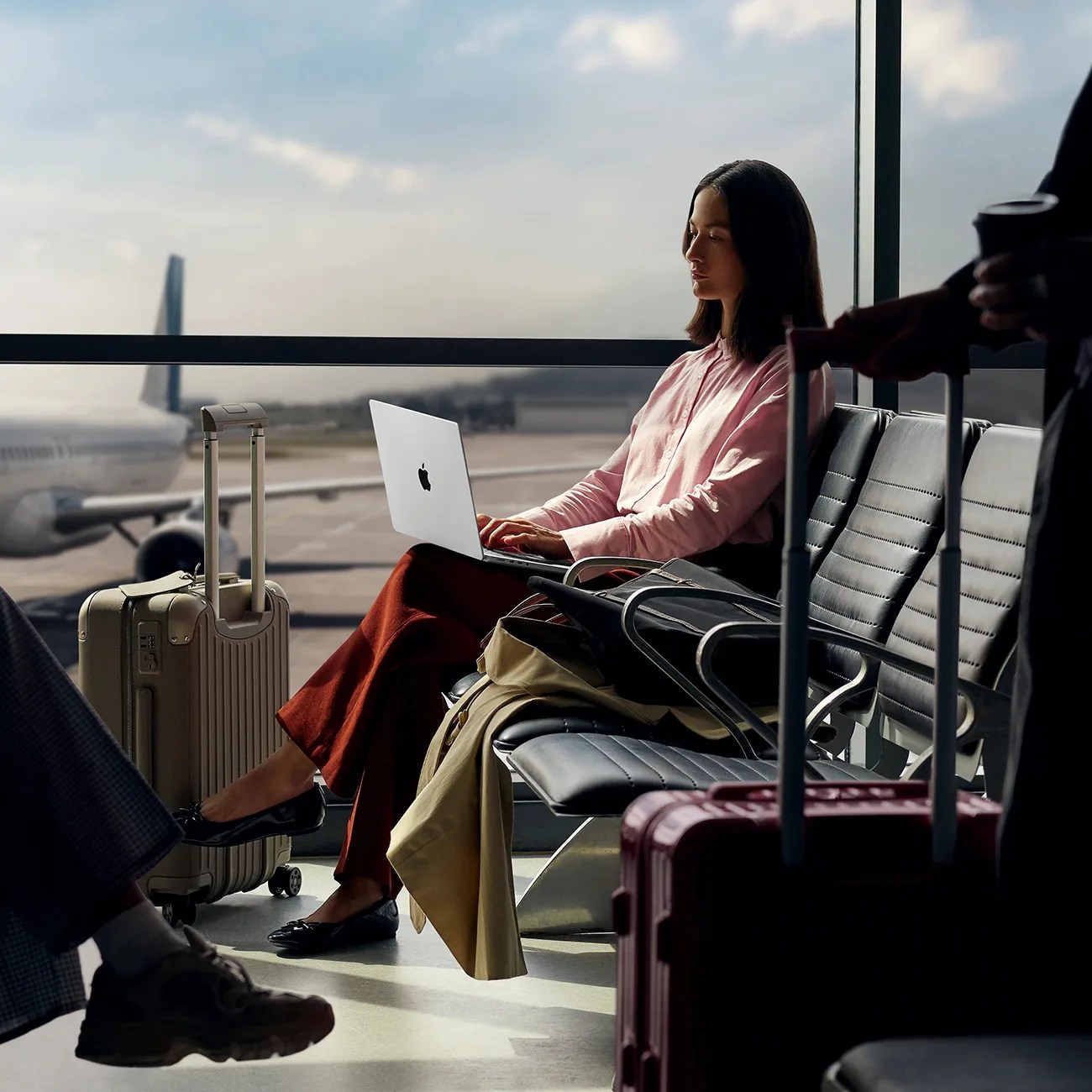 Woman in a pink shirt and rust pants working on a silver MacBook laptop while sitting in an airport lounge with beige suitcase nearby.