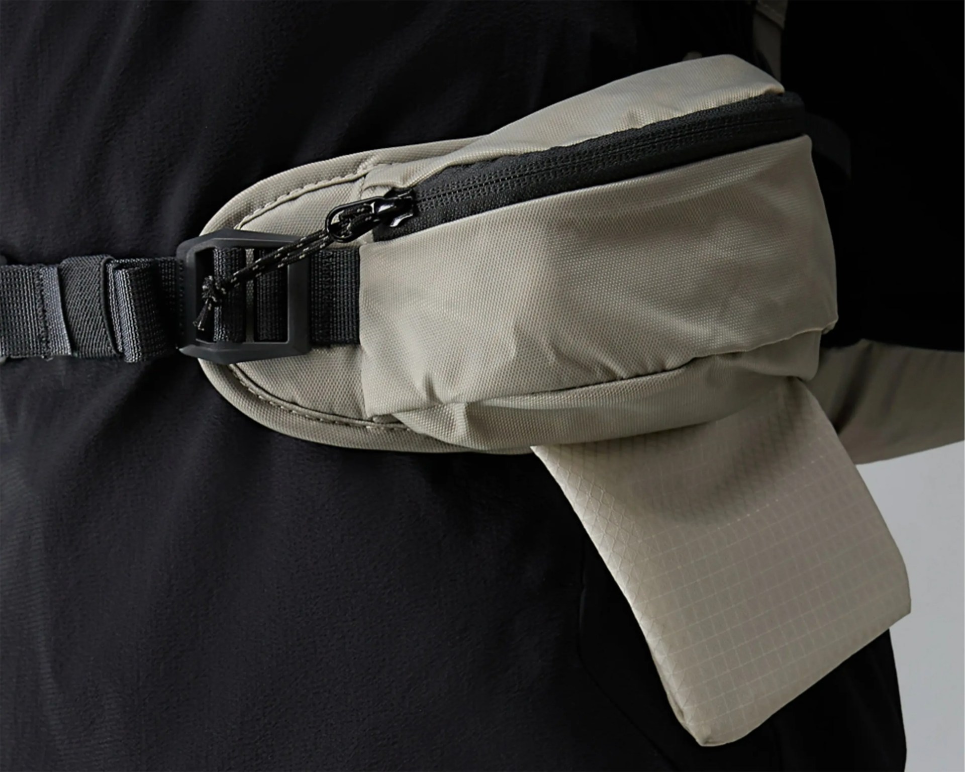 Light gray fabric waist bag with black zipper and adjustable strap worn over black clothing.