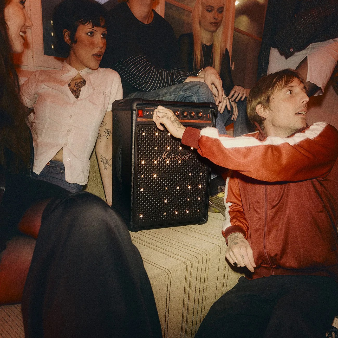Black Marshall speaker with illuminated front panel surrounded by people sitting on a couch.