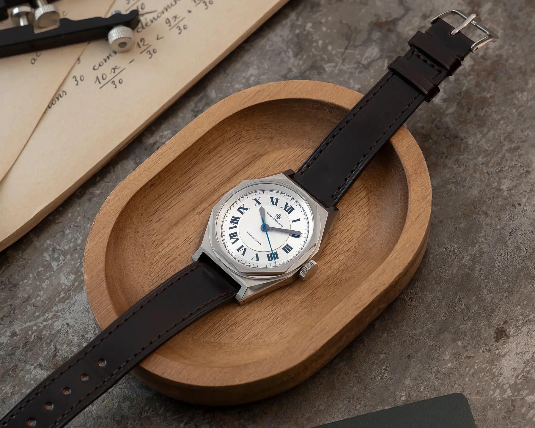 Silver wristwatch with white dial, blue Roman numerals, and black leather strap resting in a wooden dish.