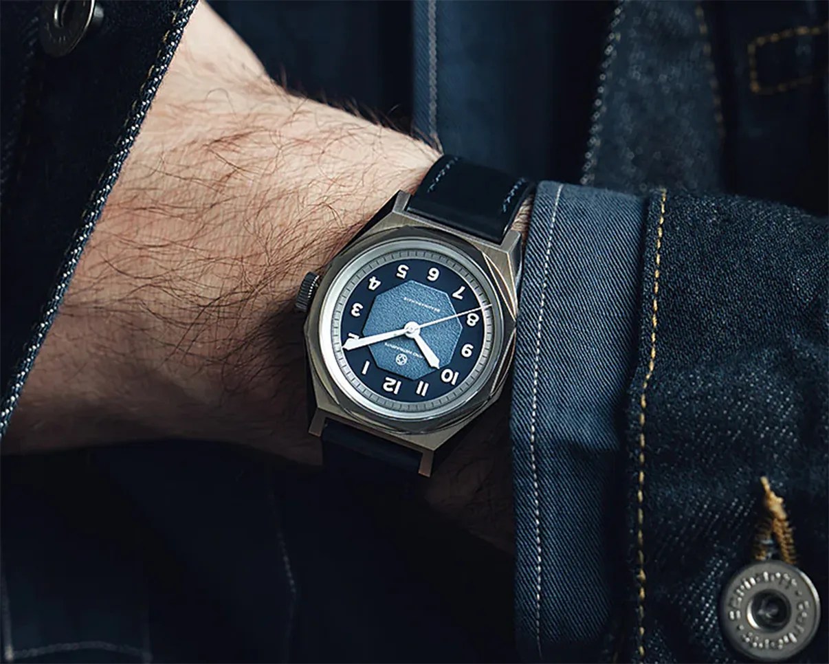 Wristwatch with a blue dial, white numerals, and a black leather strap worn on a wrist with a denim jacket.