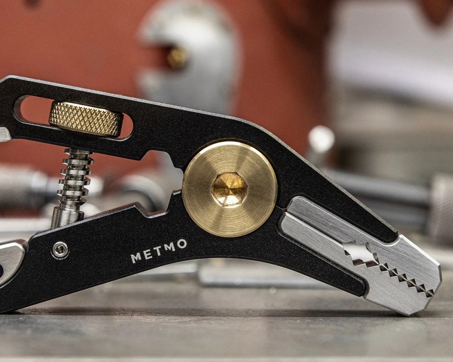Close-up of a black and silver METMO multi-tool with brass adjustment knob and pivot screw on a workbench.