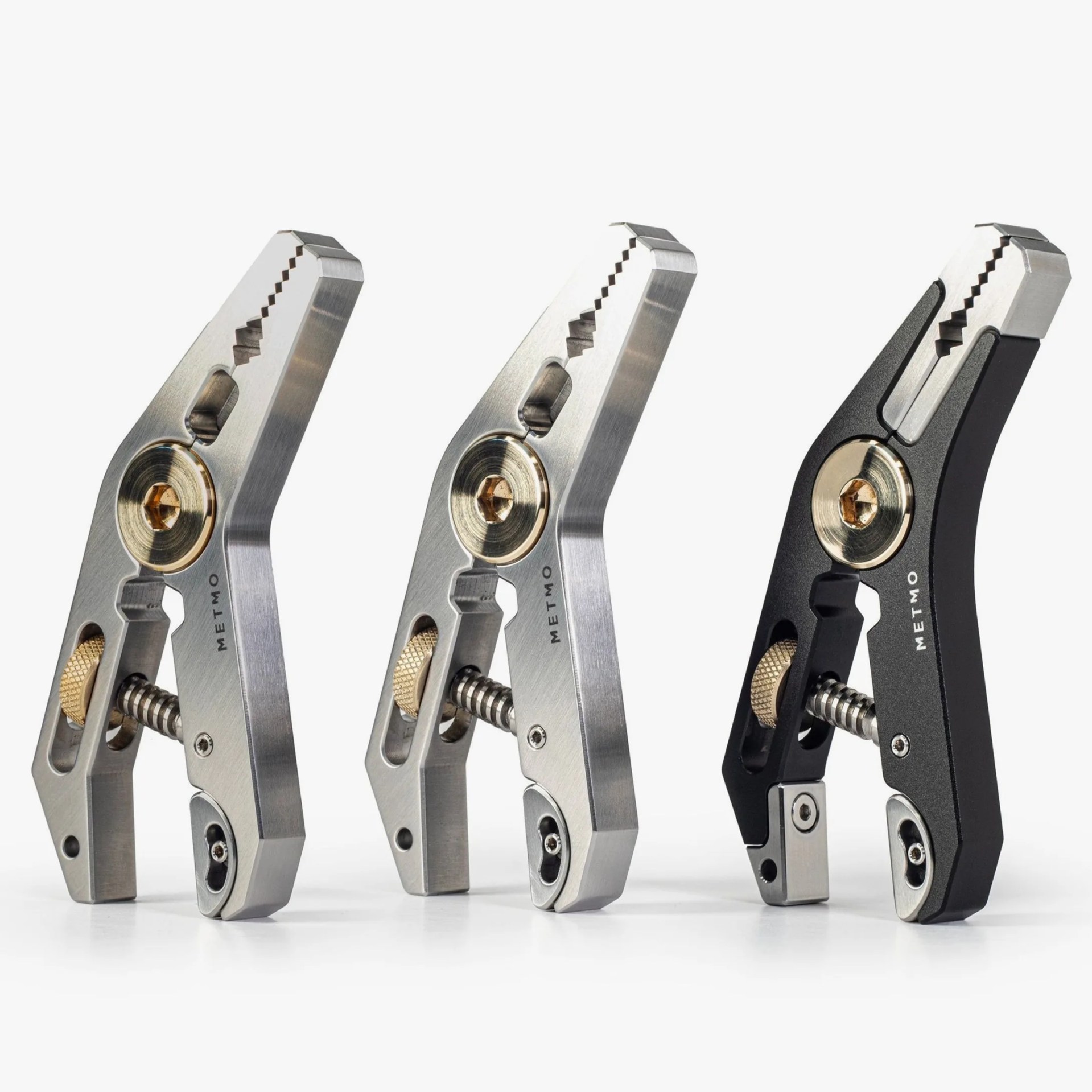 Three metal clamps with gold-colored adjustment screws, two in silver finish and one in black, standing upright on a white surface.