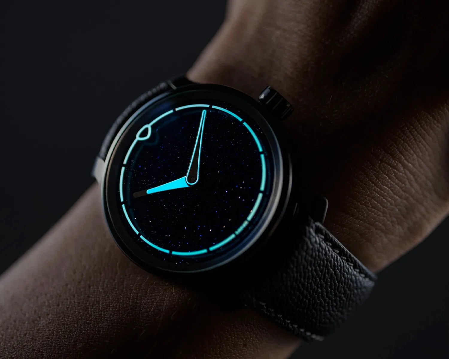 Black wristwatch with glowing blue hands and markers on a starry-patterned dial, worn on a wrist.