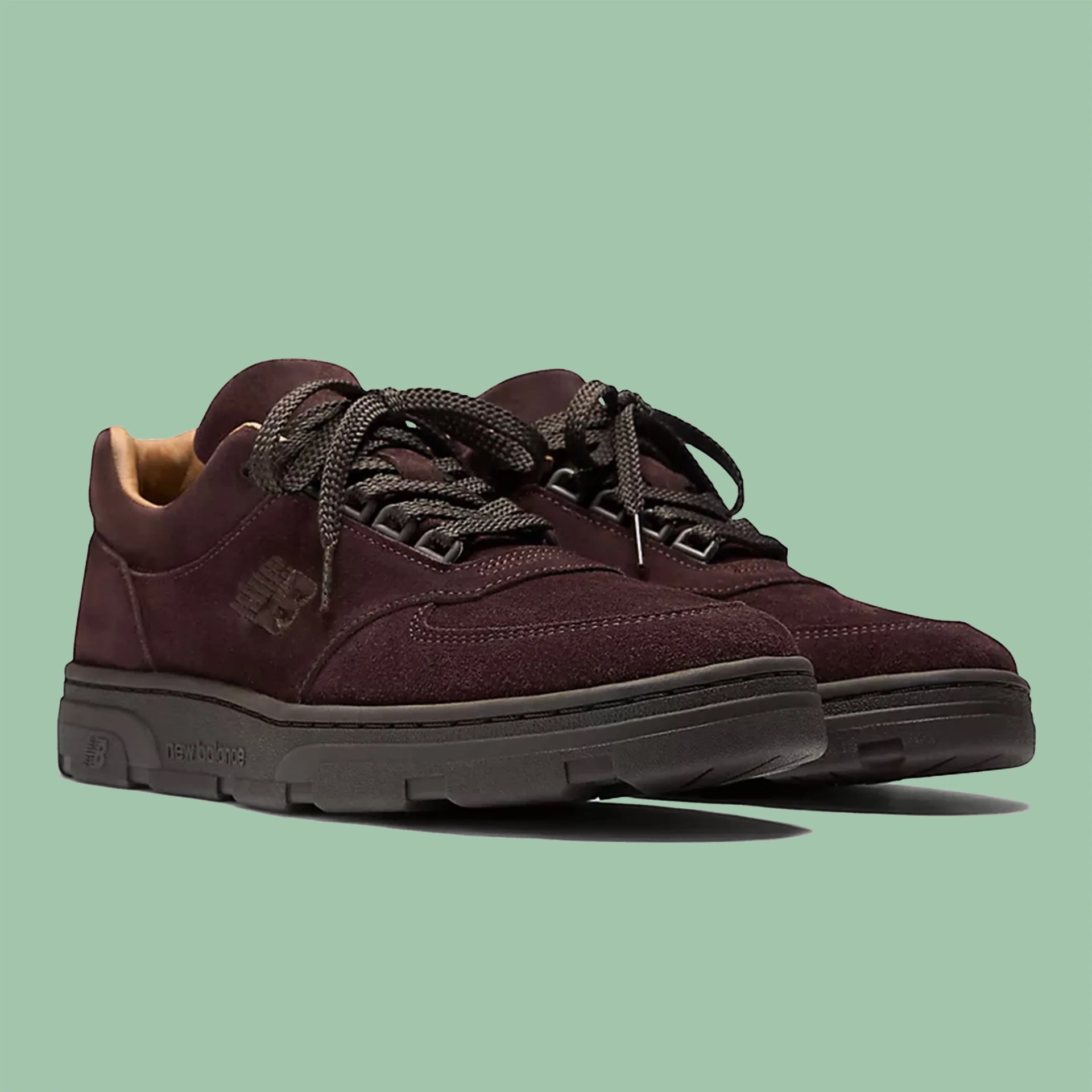 Pair of dark brown suede New Balance sneakers with black soles and thick laces on a green background.